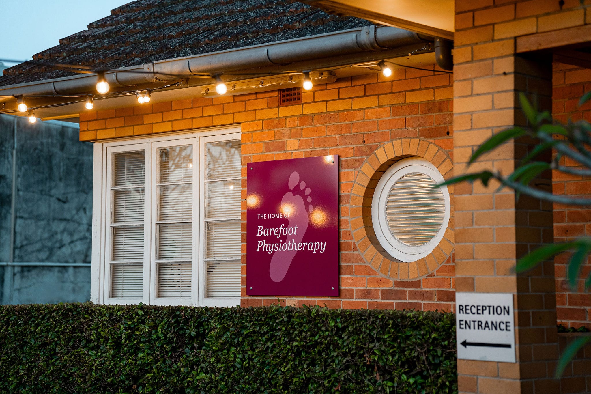 Barefoot Physiotherapy sign on red-brick clinic at dusk with string lights and “Reception Entrance” sign, Brisbane.